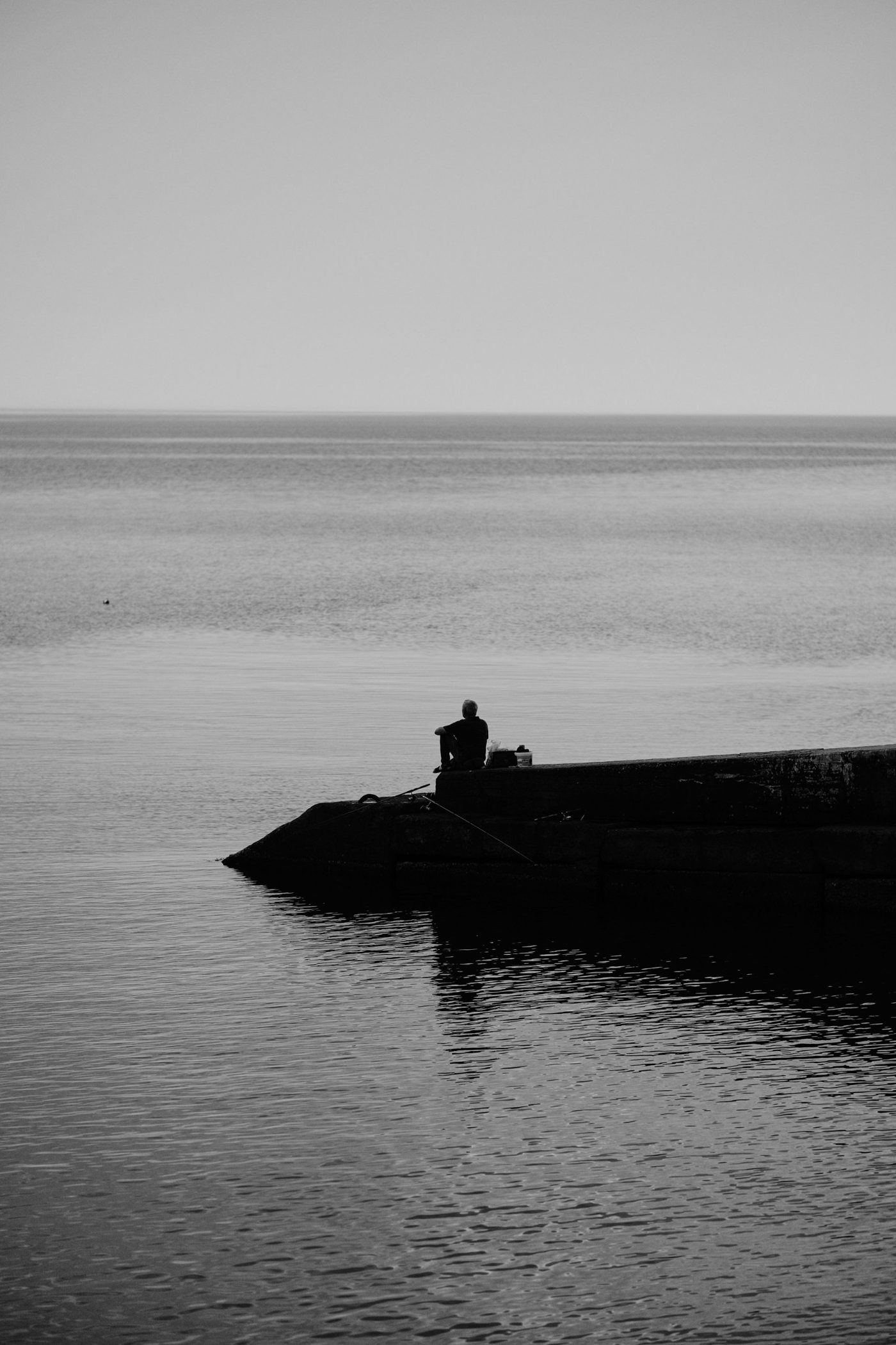 A lone man sitting at the end of a stone pier, fishing, looking out over calm open water