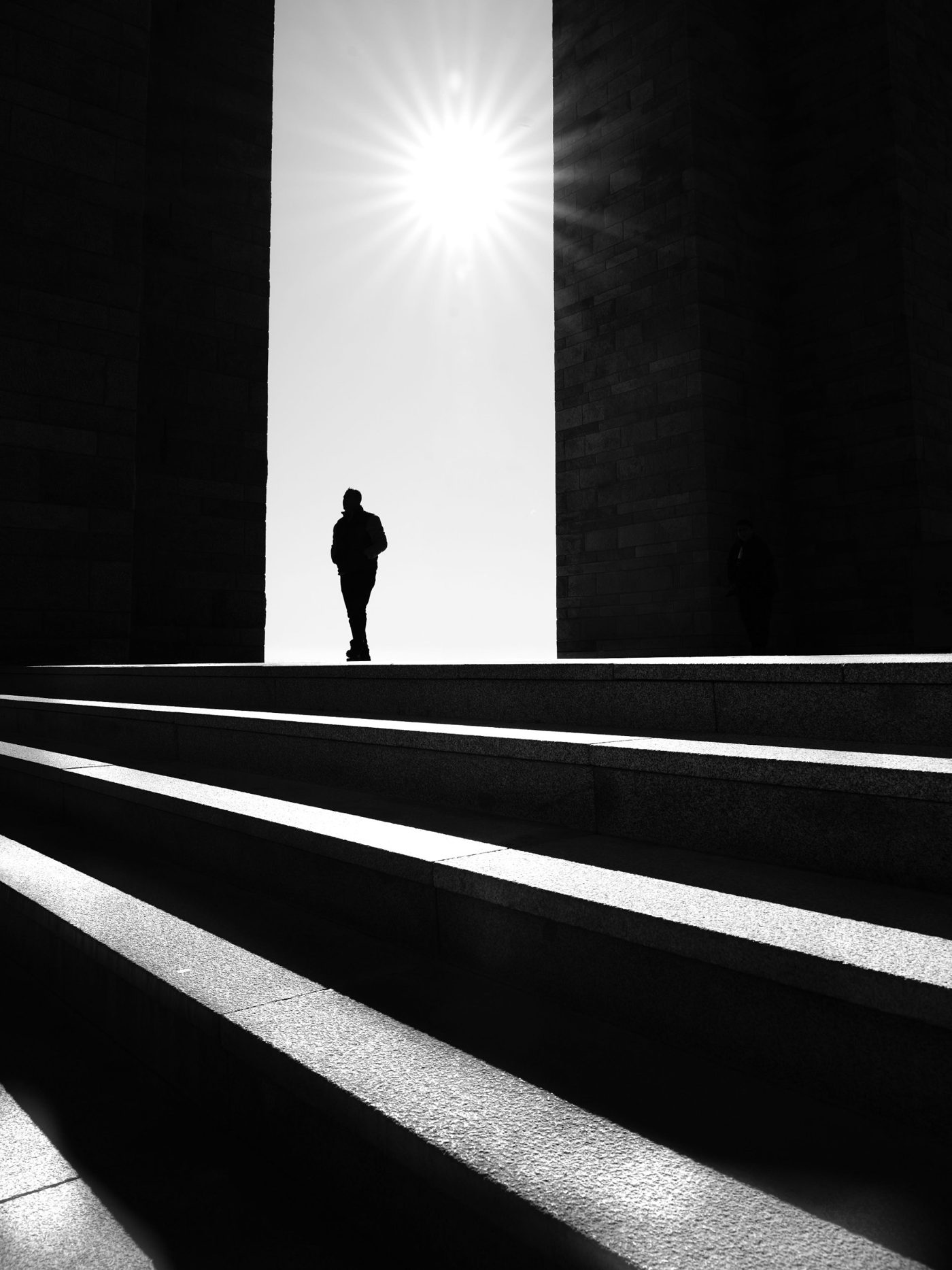 A lone man walking out from between dark stone pillars into bright sunlight, with steps in the foreground