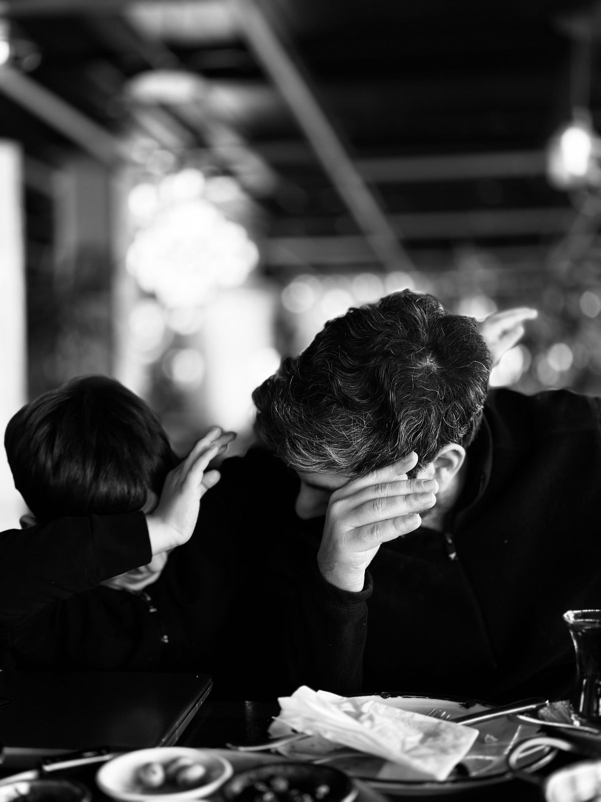 A father at a café table with his head in his hand while his young son leans against his shoulder